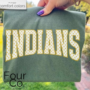 May include: A green sweatshirt with the word "INDIANS" in large, yellow polka-dotted letters. The sweatshirt is held up by a hand with purple nail polish. The brand name "Four & Co." is printed at the bottom.