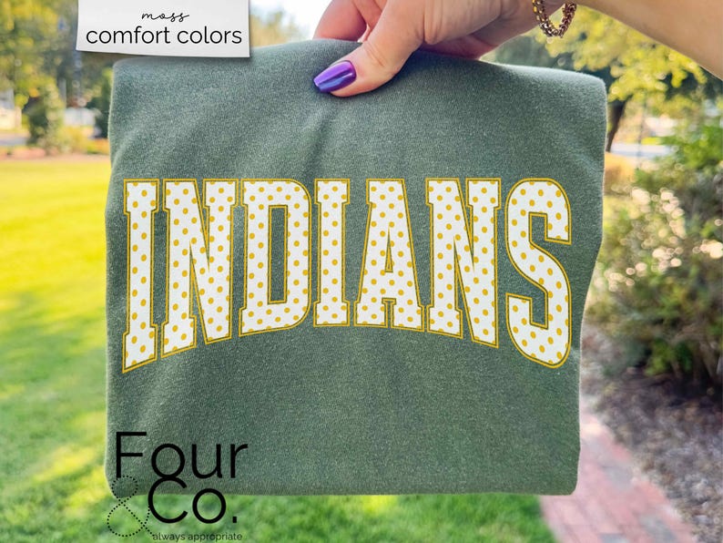 May include: A green sweatshirt with the word "INDIANS" in large, yellow polka-dotted letters. The sweatshirt is held up by a hand with purple nail polish. The brand name "Four & Co." is printed at the bottom.