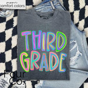 K&ouml;nnte beinhalten: Ein graues T-Shirt mit den Worten "THIRD GRADE" in gro&szlig;en, farbenfrohen, umrandeten Buchstaben. Das Shirt liegt gefaltet auf einer Jeans mit Distressed-Details. Der Hintergrund hat ein schwarz-wei&szlig;es Schachbrettmuster.