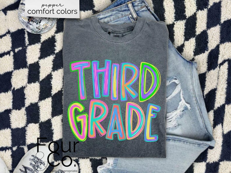 K&ouml;nnte beinhalten: Ein graues T-Shirt mit den Worten "THIRD GRADE" in gro&szlig;en, farbenfrohen, umrandeten Buchstaben. Das Shirt liegt gefaltet auf einer Jeans mit Distressed-Details. Der Hintergrund hat ein schwarz-wei&szlig;es Schachbrettmuster.
