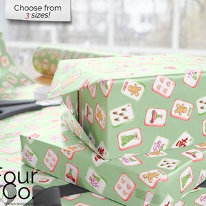 May include: Gift boxes wrapped in light green paper with a repeating pattern of playing card designs. The cards feature images of a gingerbread man, Christmas tree, and other festive icons. Scissors and a roll of wrapping paper are also visible.