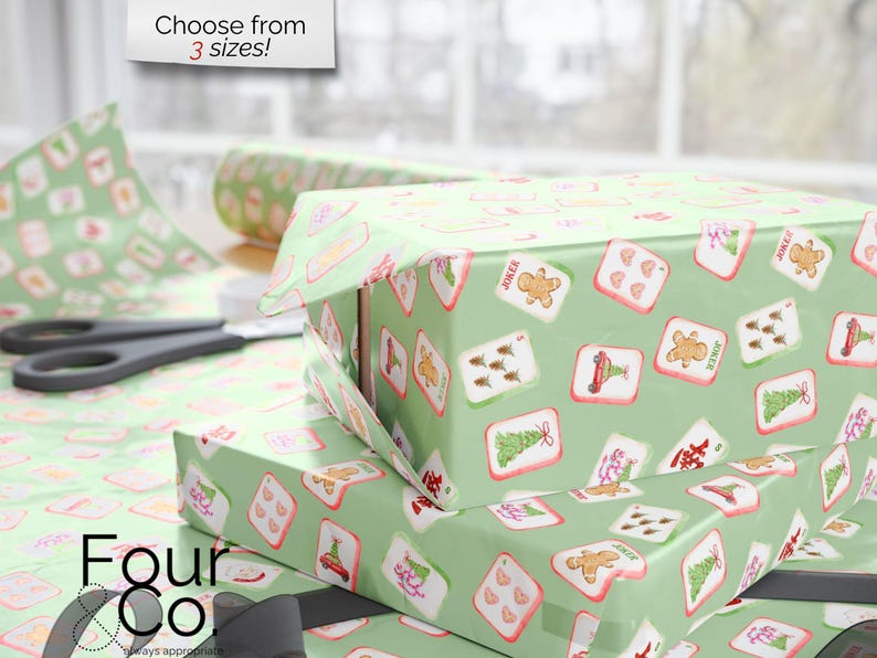 May include: Gift boxes wrapped in light green paper with a repeating pattern of playing card designs. The cards feature images of a gingerbread man, Christmas tree, and other festive icons. Scissors and a roll of wrapping paper are also visible.
