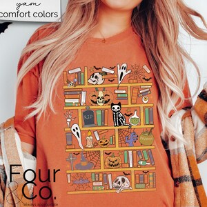 May include: A burnt orange t-shirt with a graphic of a bookshelf filled with spooky Halloween themed items, including books, pumpkins, ghosts, a skeleton cat, and a potion bottle. The text "Four & Co." is visible in the bottom left corner of the image.