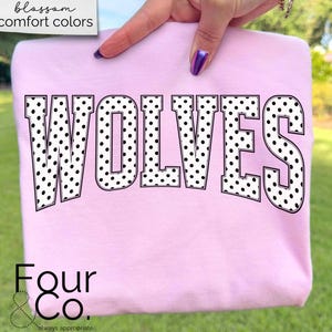 May include: A light pink sweatshirt with the word "WOLVES" in large, black and white polka dot letters. The sweatshirt is held up by a person's hand. The text "blossom comfort colors" is in the upper left corner. The brand name "Four & Co." is at the bottom.