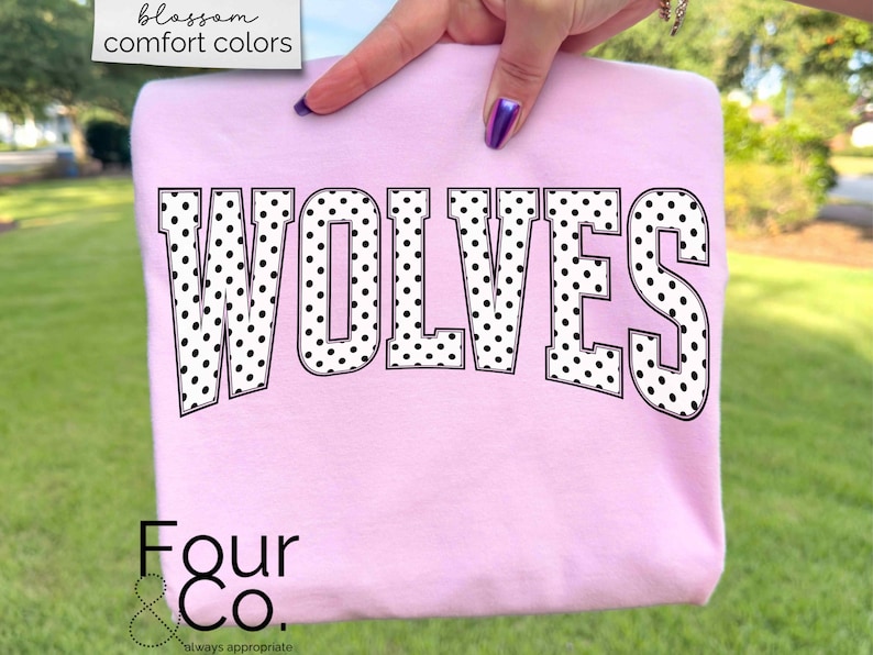 May include: A light pink sweatshirt with the word "WOLVES" in large, black and white polka dot letters. The sweatshirt is held up by a person's hand. The text "blossom comfort colors" is in the upper left corner. The brand name "Four & Co." is at the bottom.