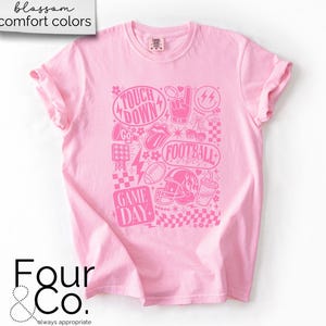 May include: Pink t-shirt with a pink graphic design featuring football-themed elements like a helmet, football, and the words "Touchdown" and "Game Day." The shirt has rolled-up sleeves and the text "blossom comfort colors" at the top.