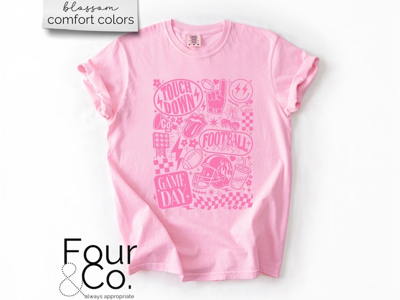 May include: Pink t-shirt with a pink graphic design featuring football-themed elements like a helmet, football, and the words "Touchdown" and "Game Day." The shirt has rolled-up sleeves and the text "blossom comfort colors" at the top.