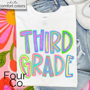 K&ouml;nnte beinhalten: Wei&szlig;es T-Shirt mit den Worten "THIRD GRADE" in bunten Buchstaben. Das Shirt wird auf einer Jeans und einer wei&szlig;en, flauschigen Oberfl&auml;che pr&auml;sentiert. Das Shirt ist von Four & Co.