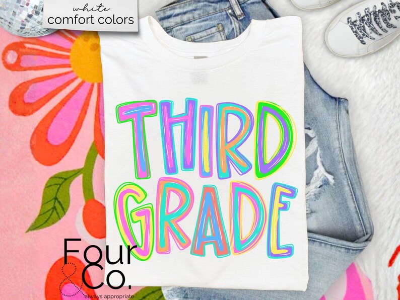 K&ouml;nnte beinhalten: Wei&szlig;es T-Shirt mit den Worten "THIRD GRADE" in bunten Buchstaben. Das Shirt wird auf einer Jeans und einer wei&szlig;en, flauschigen Oberfl&auml;che pr&auml;sentiert. Das Shirt ist von Four & Co.