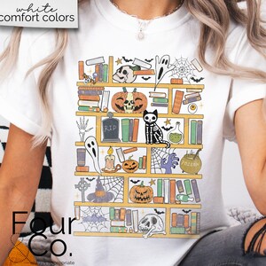 May include: A white t-shirt with a graphic of a bookshelf filled with spooky Halloween themed items, including books, pumpkins, ghosts, and a skeleton cat. The text "Four Co." is visible at the bottom of the image.
