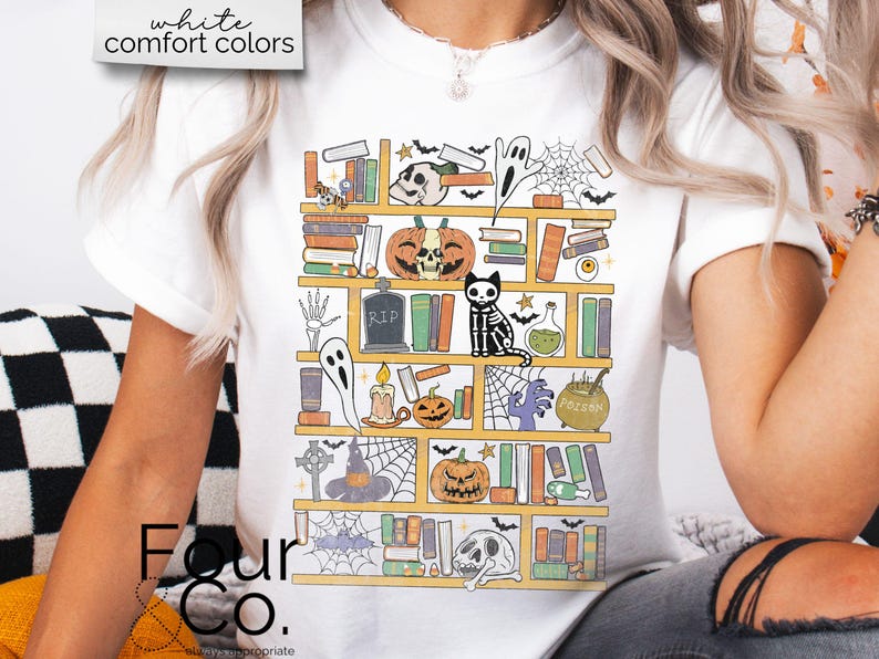 May include: A white t-shirt with a graphic of a bookshelf filled with spooky Halloween themed items, including books, pumpkins, ghosts, and a skeleton cat. The text "Four Co." is visible at the bottom of the image.