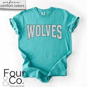 May include: A turquoise t-shirt with the word "WOLVES" in white letters with black polka dots. The shirt has rolled-up sleeves and the text "seafoam comfort colors" is in the upper left corner. The brand logo "Four & Co." is in the lower left corner.