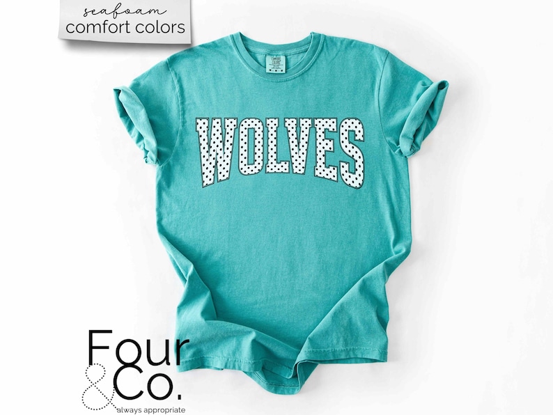 May include: A turquoise t-shirt with the word "WOLVES" in white letters with black polka dots. The shirt has rolled-up sleeves and the text "seafoam comfort colors" is in the upper left corner. The brand logo "Four & Co." is in the lower left corner.