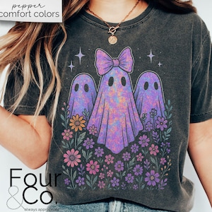Puede incluir: Camiseta gris oscuro con un dise&ntilde;o de tres fantasmas morados con un lazo, rodeados de flores coloridas. El texto "pepper comfort colors" est&aacute; en la parte superior y "Four & Co." en la inferior.