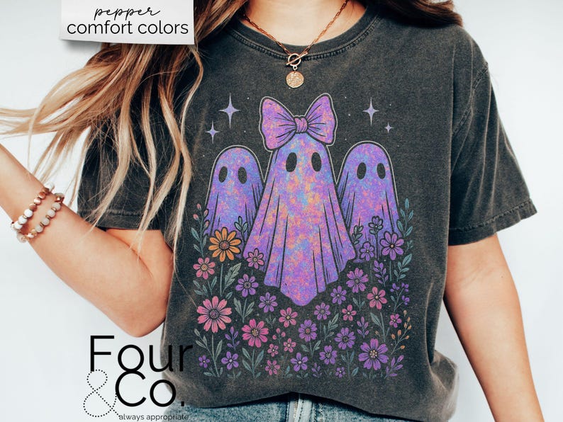 Puede incluir: Camiseta gris oscuro con un dise&ntilde;o de tres fantasmas morados con un lazo, rodeados de flores coloridas. El texto "pepper comfort colors" est&aacute; en la parte superior y "Four & Co." en la inferior.