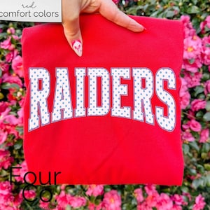 May include: A red sweatshirt with the word "RAIDERS" in white block letters with blue polka dots. The words "red comfort colors" are in a white box in the upper left corner. The sweatshirt is held up by a hand.