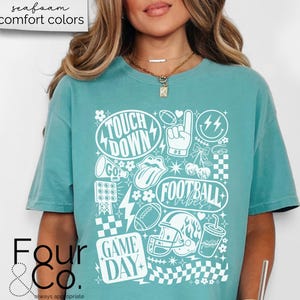 May include: A teal-colored t-shirt with a white graphic design featuring football-themed elements. The design includes the words "Touch Down", "Football Vibes", and "Game Day", along with footballs, a helmet, and other related icons. The shirt is part of the "comfort colors" collection.