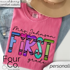 May include: Pink t-shirt with the text "Mrs. Johnson First Grade" in black lettering. The word "First" is made up of colorful crayon-shaped letters. The text "Four & Co. always appropriate" is at the bottom of the shirt.