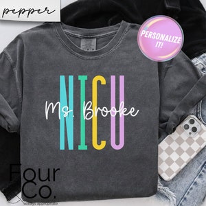 Könnte beinhalten: Dunkelgraues Langarmshirt mit dem Wort "NICU" in Türkis, Gelb und Lila sowie dem Namen "Ms. Brooke" in Weiß. Das Shirt ist gefaltet, mit einem Telefon und Jeans im Hintergrund. Das Wort "pepper" befindet sich in der oberen linken Ecke.