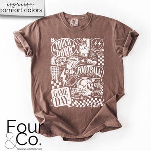 May include: Espresso-colored t-shirt with a white graphic design featuring football-themed elements. The design includes the words "Touchdown," "Football Vibes," and "Game Day," along with footballs, a helmet, and a hand signal. The shirt has a crew neck and short sleeves.