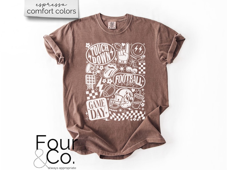 May include: Espresso-colored t-shirt with a white graphic design featuring football-themed elements. The design includes the words "Touchdown," "Football Vibes," and "Game Day," along with footballs, a helmet, and a hand signal. The shirt has a crew neck and short sleeves.