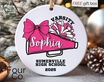 May include: A white ceramic ornament with a pink megaphone, bow, and pom-poms design. The ornament features the name "Sophia", "Varsity", "Summerville High School", and the year "2025". A gold string is attached for hanging.