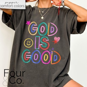 以下が含まれることがあります： カラフルな文字で「GOD IS GOOD」と書かれた、濃いグレーのTシャツ。デザインにはスマイリーフェイス、ハート、星が含まれています。ゆったりとしたフィット感で、半袖です。