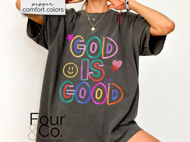 以下が含まれることがあります： カラフルな文字で「GOD IS GOOD」と書かれた、濃いグレーのTシャツ。デザインにはスマイリーフェイス、ハート、星が含まれています。ゆったりとしたフィット感で、半袖です。