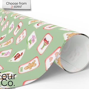 May include: A roll of wrapping paper with a light green background and a repeating pattern of Christmas-themed playing cards. The cards feature gingerbread men, Christmas trees, and other festive designs. The text "Choose from 3 sizes!" is visible.