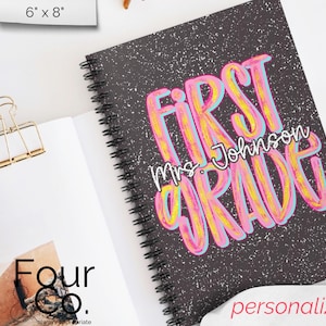 May include: A spiral-bound notebook with a black cover featuring the words "FIRST GRADE" in large, colourful letters. The name "Mrs. Johnson" is written below. The notebook is open to a blank page, with a gold binder clip.