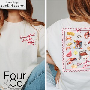 May include: A white t-shirt with a red "Crawfish Season" graphic on the front and a red stamp-like graphic with a variety of crawfish-related imagery on the back. The shirt is being modeled by a person wearing blue jeans.