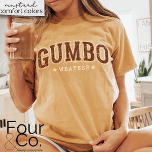May include: A mustard yellow t-shirt with the text "GUMBO WEATHER" in brown letters with a vintage distressed look. The shirt is worn by a person holding a glass of iced coffee.