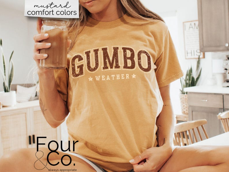 May include: A mustard yellow t-shirt with the text "GUMBO WEATHER" in brown letters with a vintage distressed look. The shirt is worn by a person holding a glass of iced coffee.