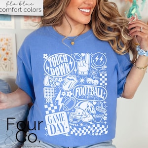 May include: A blue t-shirt with a white graphic design featuring football-themed elements. The design includes the words "Touch Down", "Football Vibes", and "Game Day", along with footballs, a helmet, and a foam finger. The shirt also has the text "flo blue comfort colors".