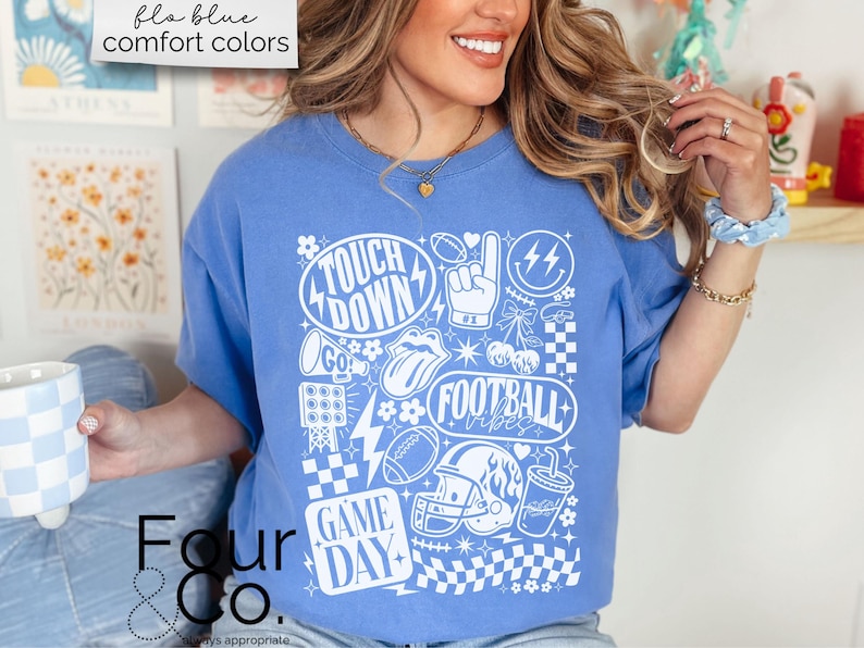 May include: A blue t-shirt with a white graphic design featuring football-themed elements. The design includes the words "Touch Down", "Football Vibes", and "Game Day", along with footballs, a helmet, and a foam finger. The shirt also has the text "flo blue comfort colors".