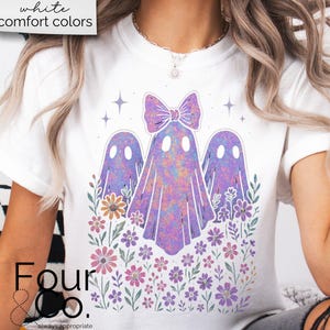 Puede incluir: Camiseta blanca con un dise&ntilde;o caprichoso de tres fantasmas morados con un lazo, rodeados de flores y estrellas coloridas. El texto "white comfort colors" est&aacute; en la parte superior. La marca "Four & Co." est&aacute; en la parte inferior.