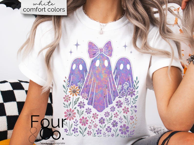 Puede incluir: Camiseta blanca con un dise&ntilde;o caprichoso de tres fantasmas morados con un lazo, rodeados de flores y estrellas coloridas. El texto "white comfort colors" est&aacute; en la parte superior. La marca "Four & Co." est&aacute; en la parte inferior.