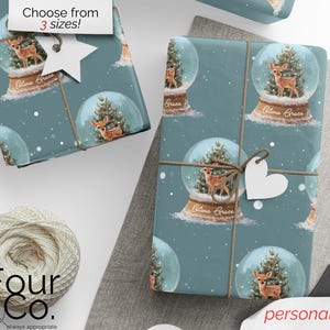 May include: Gift wrap featuring a repeating pattern of snow globes with a deer and Christmas tree inside. The background is a teal color. The words "Olivia Grace" are on the snow globe base. A white heart-shaped tag is tied to the wrapped package.
