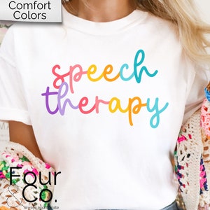 May include: White t-shirt with the text "speech therapy" in rainbow colors. The text is in a handwritten font. The shirt is being worn by a person with long blonde hair. The person is wearing a crocheted cardigan.