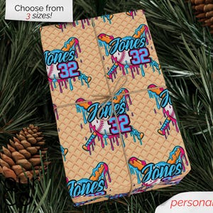 May include: Wrapped gift with a baseball-themed design. The wrapping paper features a repeating pattern of baseballs, bats, and the name "Jones 32" in blue, orange, and pink. The gift is tied with twine and set against a backdrop of pine needles and pine cones.