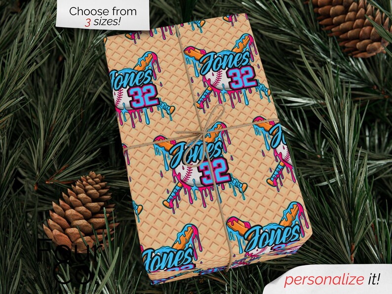May include: Wrapped gift with a baseball-themed design. The wrapping paper features a repeating pattern of baseballs, bats, and the name "Jones 32" in blue, orange, and pink. The gift is tied with twine and set against a backdrop of pine needles and pine cones.