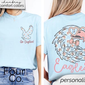 Pu&ograve; includere: T-shirt azzurra con un motivo a forma di aquila. La parte anteriore presenta una piccola aquila con un motivo floreale e la scritta "Go Eagles!". Il retro mostra un'aquila pi&ugrave; grande con occhiali a forma di cuore e un papillon, con la parola "Eagles" sotto.