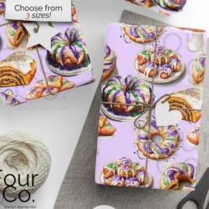 May include: Two rolls of wrapping paper with a purple background and a watercolor pattern of king cakes. The wrapping paper is tied with twine and has a heart-shaped tag. The text "Choose from 3 sizes!" is visible.