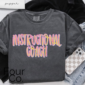 May include: A dark gray long-sleeve shirt with the words "INSTRUCTIONAL COACH" in a colorful, bubble-style font. The shirt is paired with jeans and a phone case with a checkered pattern.