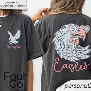Pu&ograve; includere: T-shirt grigio scuro con un design floreale di aquila con occhiali da sole a forma di cuore e un fiocco. La parte anteriore ha una grafica di aquila pi&ugrave; piccola e le parole "Go Eagles!". La parte posteriore della maglietta ha la parola "Eagles" in corsivo.