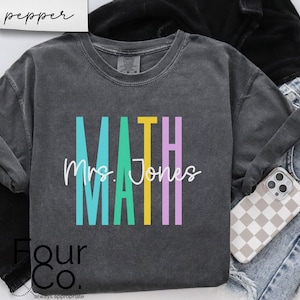 May include: A dark gray t-shirt with the word "MATH" in colorful vertical stripes and the text "Mrs. Jones" in white script below. The shirt is made of a soft, comfortable material and is perfect for teachers.
