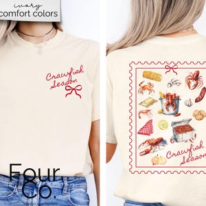May include: A white t-shirt with the text "Crawfish Season" in red script on the front and a red bow. The back of the shirt has a red and white stamp design with the text "Crawfish Season" and illustrations of crawfish, corn, garlic, and other ingredients.