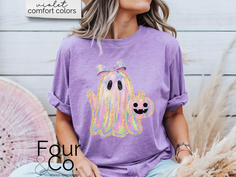 May include: A purple t-shirt with a holographic ghost holding a pumpkin. The ghost has a pink bow on its head. The text "violet comfort colors" is visible at the top of the image.
