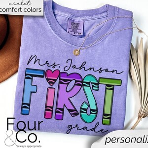May include: Purple t-shirt with the text "Mrs. Johnson First Grade" in black lettering. The word "First" is spelled out with colorful crayon-like letters. The text "Four & Co. always appropriate" is at the bottom of the shirt.