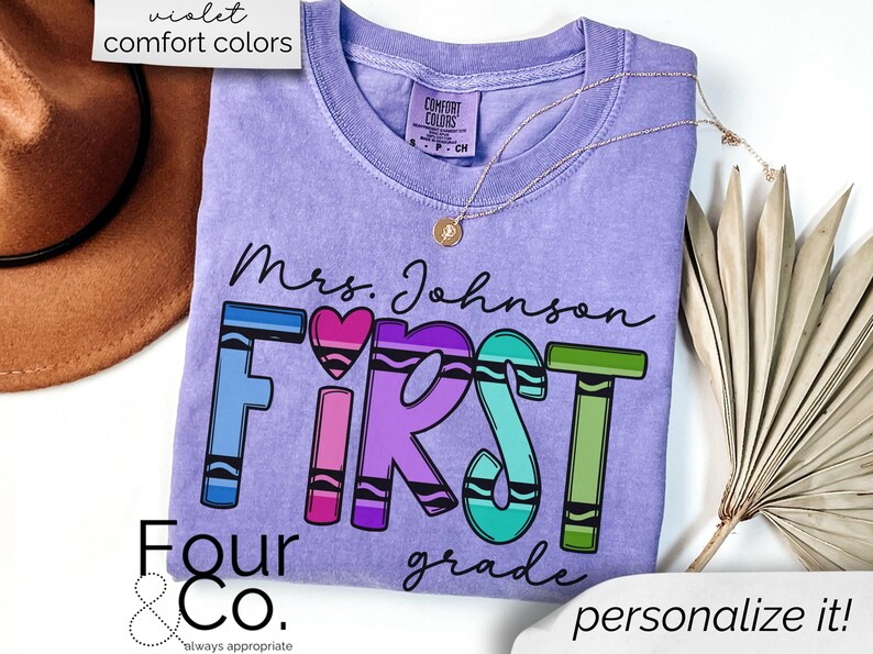 May include: Purple t-shirt with the text "Mrs. Johnson First Grade" in black lettering. The word "First" is spelled out with colorful crayon-like letters. The text "Four & Co. always appropriate" is at the bottom of the shirt.